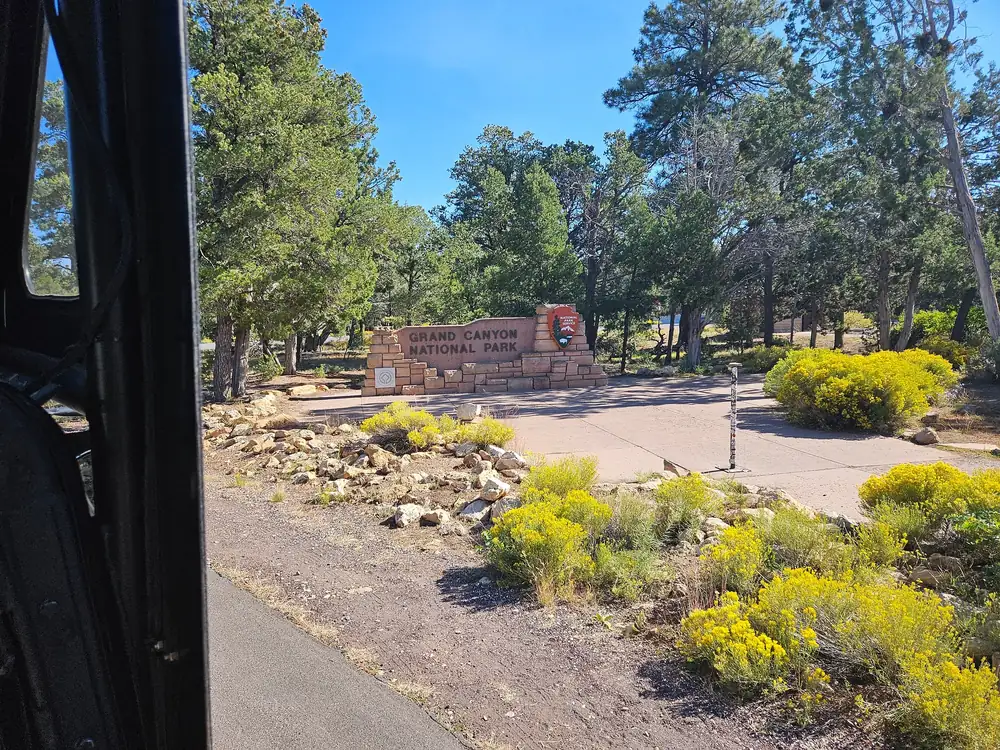 Das Schild des Grand-Canyon-Nationalparks zwischen Kiefern und gelben Sträuchern, aus einem Tourfahrzeug gesehen