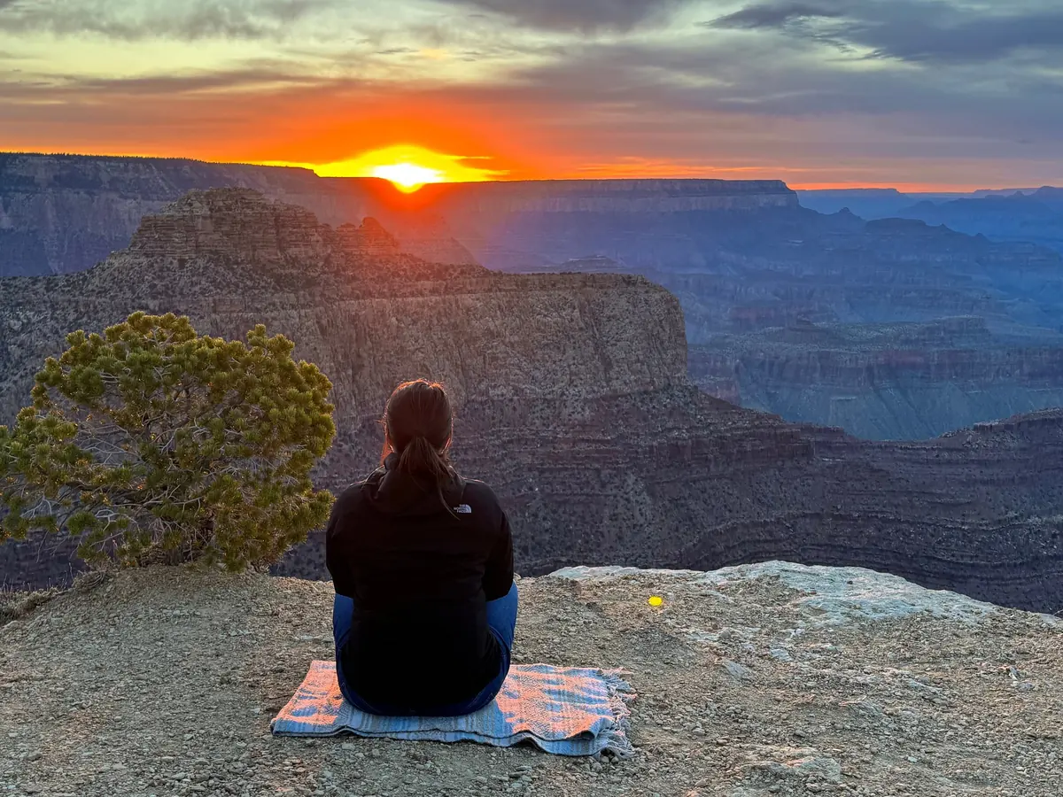 A visitor sitting on a blanket at the rim watching the sun set over the canyon